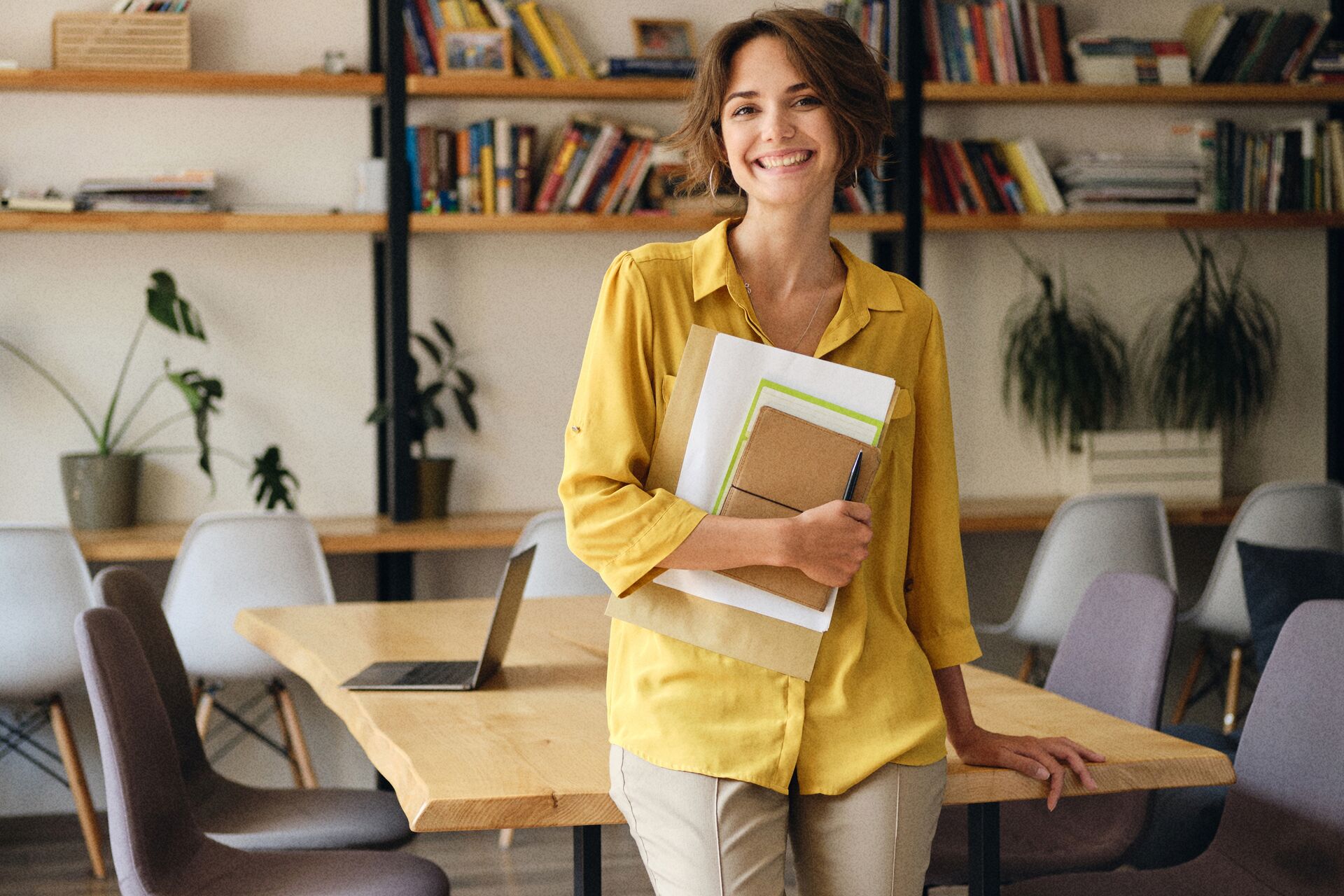Young cheerful woman in yellow shirt leaning on desk with notepad and papers in hand joyfully looking in camera in modern office